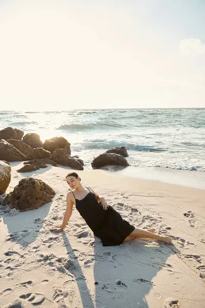 A young woman in a black sundress lounges on the beach near the ocean at sunset. — Stock Photo