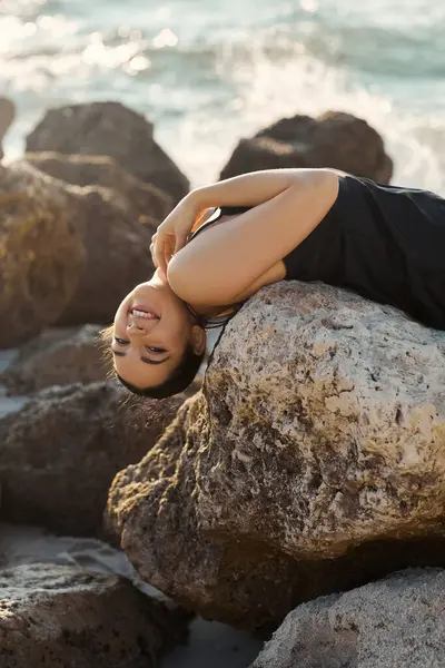 A young woman in a black sundress smiles as she lies on a large rock near the ocean on Miami Beach. — Stock Photo