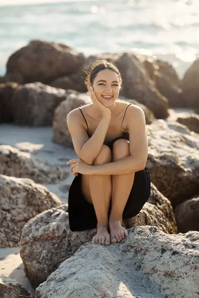 Una giovane donna in un sundress nero sorride luminosa mentre si siede su una roccia vicino all'oceano a Miami Beach. — Foto stock