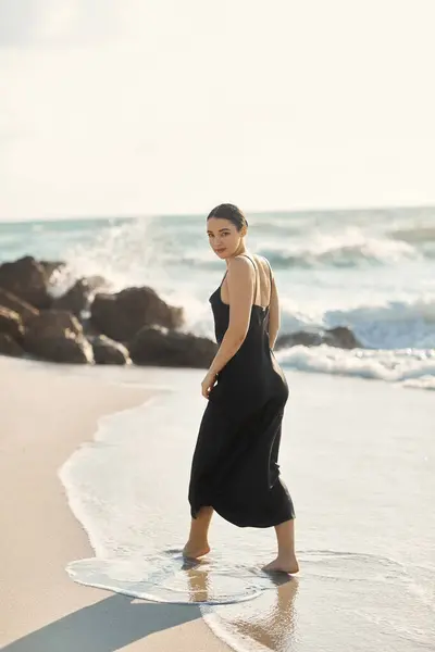 A young woman in a black sundress walks along the shore of Miami Beach, Florida, as waves gently lap at her feet. — Stock Photo