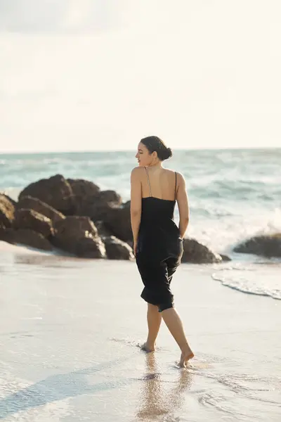A woman in a black sundress walks barefoot along the shoreline of Miami Beach, the setting sun casting a warm glow over the waves. — Stock Photo