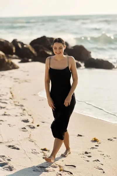 A young woman in a black sundress walks barefoot along the shore of Miami Beach, Florida, with the ocean in the background. — Stock Photo