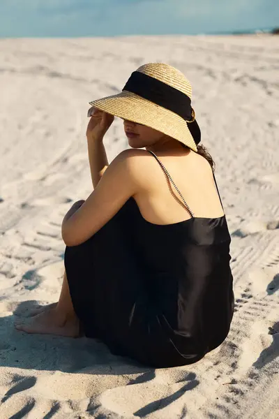 Una giovane donna con un sundress nero e un cappello di paglia siede sulla sabbia, il suo sguardo perso all'orizzonte. — Foto stock