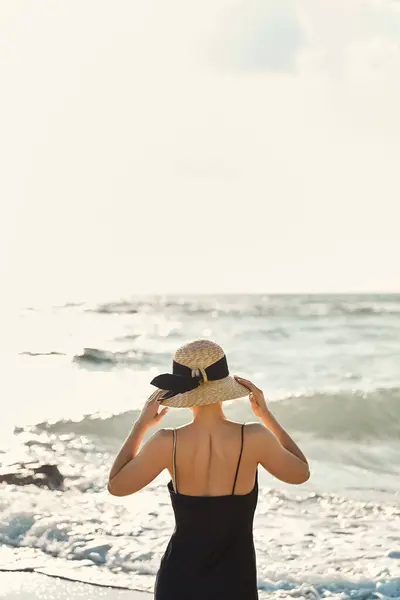 A young woman in a black sundress and a straw hat stands on the beach, facing the ocean with her back to the camera. — Stock Photo