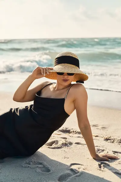 Uma jovem mulher em um vestido preto e chapéu de palha salões na costa arenosa de Miami Beach, desfrutando do sol quente e suave brisa do mar. — Fotografia de Stock