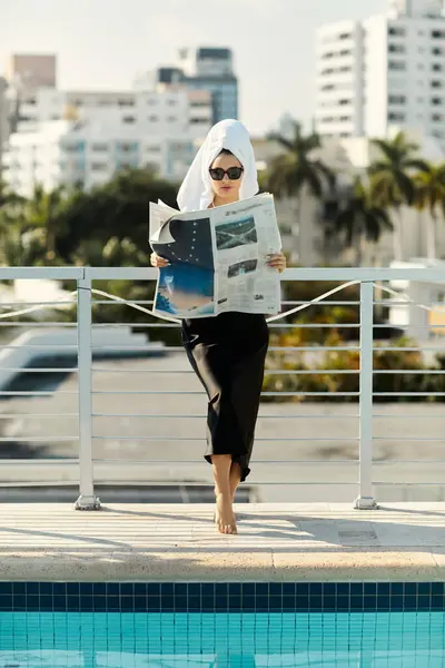 A stylish young woman, wearing sunglasses and a black slip dress, relaxes by the pool in Miami, reading a newspaper. — Stock Photo