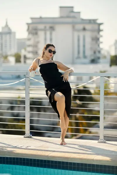 A stylish woman in a black slip dress leans on a railing by the pool, sunglasses perched on her face. — Stock Photo