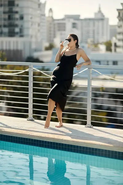 Eine elegante junge Frau im schwarzen Slip-Kleid genießt einen morgendlichen Kaffee auf einem Villenbalkon mit Blick auf einen Pool in Miami. — Stockfoto