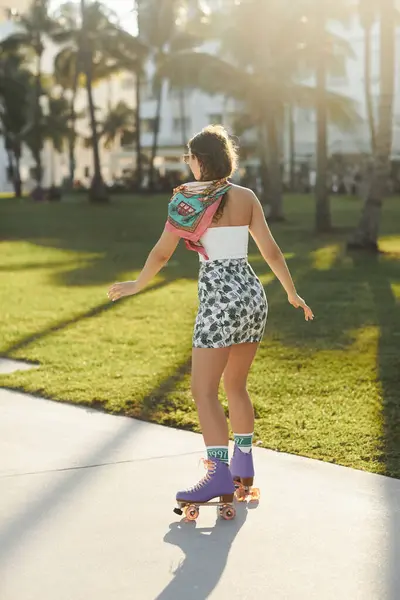 A young woman in sunglasses and stylish attire skates on a sidewalk under palm trees in Miami. — Stock Photo