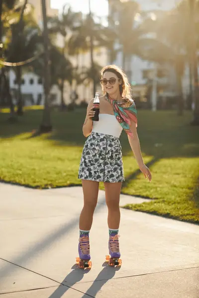 A young woman in sunglasses and a floral skirt rollerblades down a sidewalk in Miami, enjoying a cool drink on a sunny day. — Stock Photo