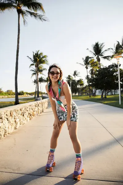 A young woman in sunglasses and a stylish outfit rollerblades along a paved pathway in Miami, palm trees surrounding her. — Stock Photo