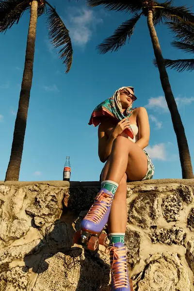 A young woman sits gracefully on a stone wall next to lush palm trees in a stylish outfit and sunglasses, enjoying a peaceful moment. — Stock Photo