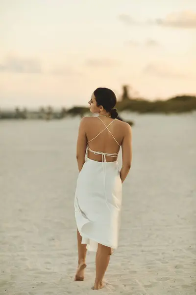 A young woman strolls barefoot along the beach in Miami, her white dress flowing behind her. — Stock Photo