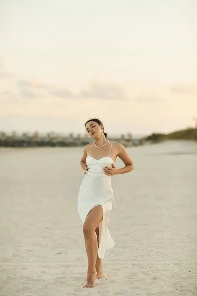 A young woman in a white dress walks barefoot on a sandy beach in Miami as the sun sets behind her. — Stock Photo