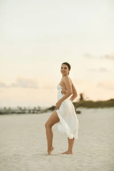 A beautiful young woman in a flowing white dress smiles as she walks along the sandy shore of Miami Beach. — Stock Photo