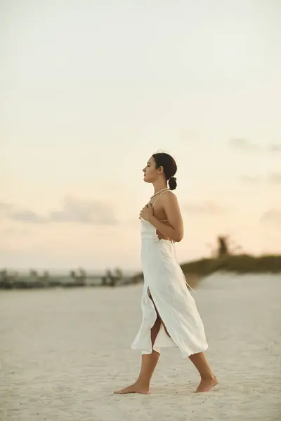 A young woman in a white dress walks barefoot on a sandy beach at sunset. — Stock Photo