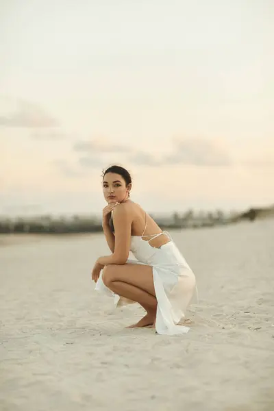 A young woman in a white dress sits on the beach in Miami, looking out at the horizon. — Stock Photo
