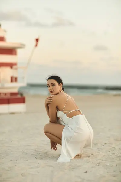 Una joven con un vestido blanco se sienta en la arena de Miami Beach, sus ojos mirando a la distancia. - foto de stock