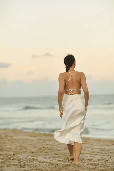 A woman in a flowing white dress walks barefoot on a sandy beach in Miami, the sun setting in the distance. — Stock Photo