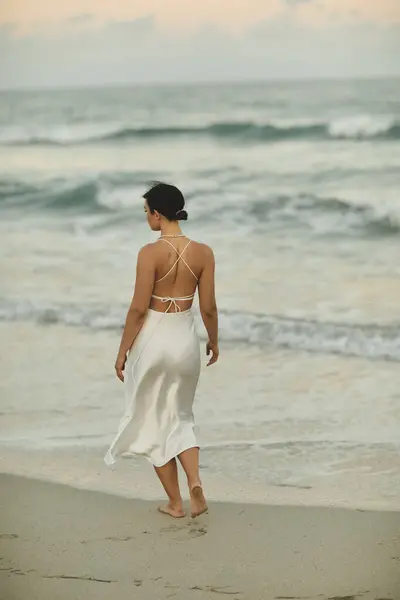 A young woman in a white dress walks barefoot along a sandy beach in Miami. — Stock Photo