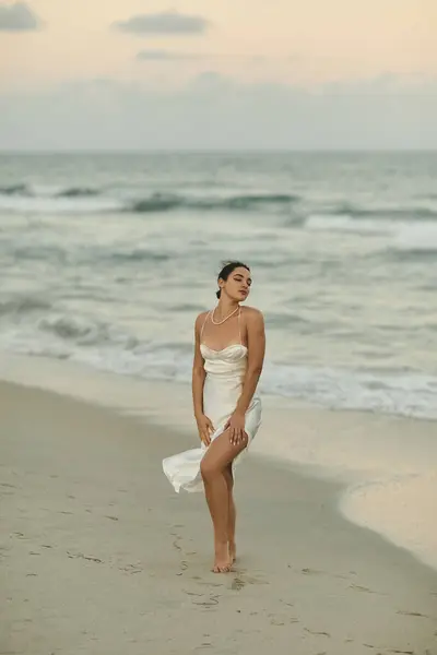 A beautiful young woman in a flowing white dress stands on a sandy beach in Miami, the setting sun casting a warm glow on the scene. — Stock Photo