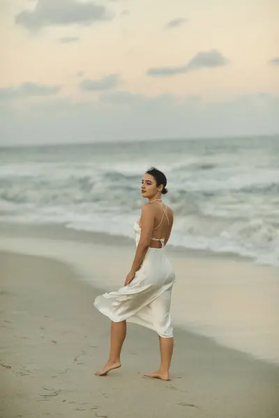 Una giovane donna cammina su una spiaggia di sabbia a Miami, indossando un abito bianco fluente, mentre il sole tramonta dietro di lei.. — Foto stock