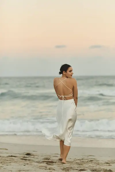 A young woman in a white dress walks along a sandy beach in Miami, the setting sun casting a warm glow on the horizon. — Stock Photo