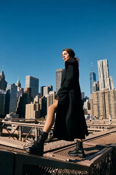 Una giovane donna con un cappotto nero attraversa il ponte di Brooklyn, guardando verso lo skyline della città. — Foto stock