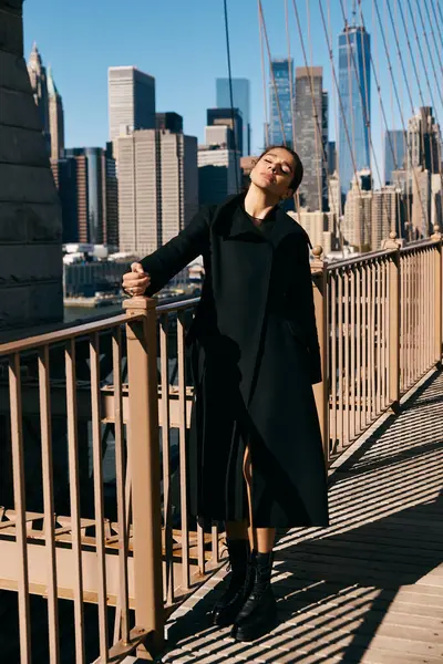 Una joven con un abrigo negro baila en un puente con vistas al horizonte de Nueva York. - foto de stock