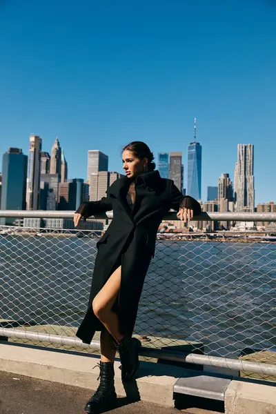 Mujer con abrigo negro posa en un muelle de Nueva York, horizonte en el fondo. - foto de stock