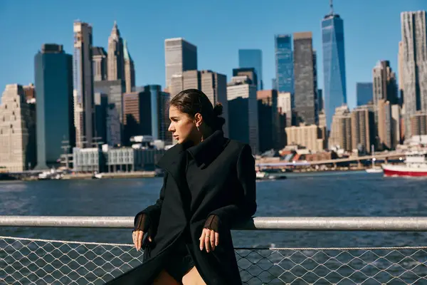 Eine junge Frau im schwarzen Mantel lehnt an einem Geländer und blickt auf die Skyline von New York City. — Stockfoto