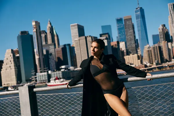 Una joven con un traje negro baila en un muelle con vistas al horizonte de la ciudad de Nueva York. - foto de stock