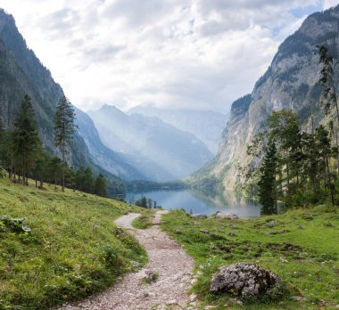 Gün batımında, kozalaklı ormanın ortasında, kayalık duvarlar arasında turkuaz suyla Obersee Gölü. Köknar ağaçları arasında bir dağ gölü manzarası. Bulutlarda parlayan güneş ışınları.