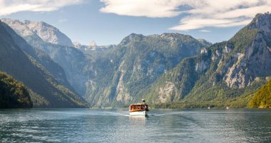 Sakin bir göl, parçalı bulutlu bir gökyüzünün altında, suyun üzerinde süzülen küçük bir kayıkla, yüksek, ormanlı dağları yansıtır. Manzara, görkemli manzara seyahat, doğa ve macera için mükemmeldir.