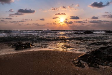 Gün batımı, bulutlardan çıkan güneş ışınları, güçlü dalgalarla gelen deniz. 