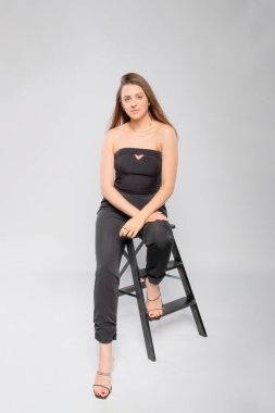  Siyah elbiseli manken stüdyoda gri arka planda oturuyor. .