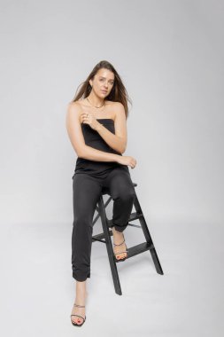 Şık siyah takım elbiseli şık bir kadın gri izole bir arka planda bir sandalyeye oturmuş poz veriyor. Çekici bir mankenin portresi.