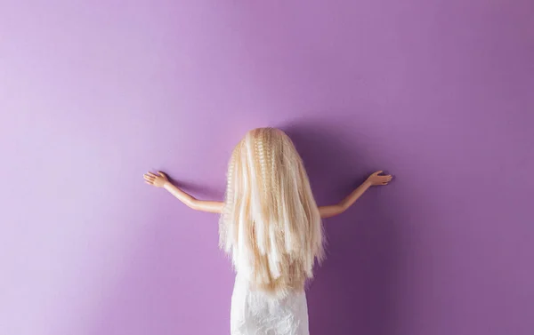 Girl doll stands alone against a purple wall with outstretched arms Minimal concept.