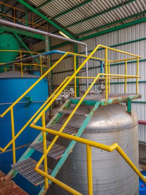 Sarı güvenlik korkuluğu ve merdiveni olan büyük bir çelik tankın olduğu sanayi sahnesi. Mavi bir tank ve çeşitli borular arka planda kıvrımlı bir çatı altında görünür.