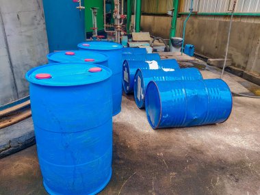 Endüstriyel ekipmanların yanında kırmızı kapaklı büyük mavi plastik fıçılar dizilmiştir.