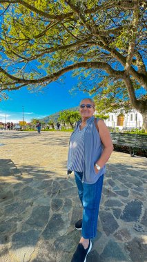 Gri saçlı, mavi gömlekli, kot pantolonlu, parktaki bir ağacın altında duran yaşlı güzel bir kadının portresi..