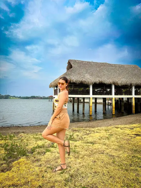 Gölün kenarında tropikal bir tatilin tadını çıkaran bir kadın. Üzerinde ay çiçekleri olan bir yaz elbisesi, canlı bir gökyüzünün altındaki sazlı çatı iskelesinin yanında..