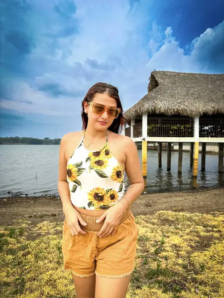 Gölün kenarında tropikal bir tatilin tadını çıkaran bir kadın. Üzerinde ay çiçekleri olan bir yaz elbisesi, canlı bir gökyüzünün altındaki sazlı çatı iskelesinin yanında..