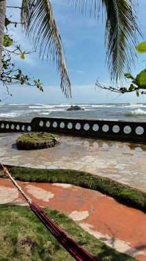 Kırmızı bir hamağı, yemyeşil yeşilliği ve kayalara çarpan dalgalarla okyanusa bakan taş bir duvarı olan sakin bir tropikal manzara. Seyahat, rahatlama ve doğa temaları için mükemmel.. 