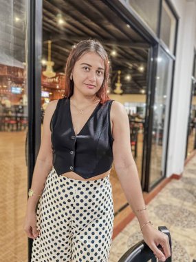 Siyah yelek ve puantiyeli şık bir kadın, geniş pencereli ve zarif dekorlu modern bir restoranda güven içinde poz veriyor. Moda, yaşam tarzı ve yemek temaları için mükemmel. Yüksek kalite, şık.