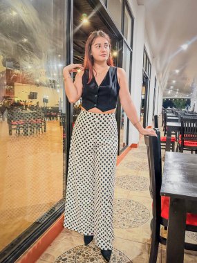 Siyah yelek ve puantiyeli şık bir kadın, geniş pencereli ve zarif dekorlu modern bir restoranda güven içinde poz veriyor. Moda, yaşam tarzı ve yemek temaları için mükemmel. Yüksek kalite, şık.