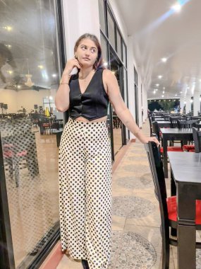 Siyah yelek ve puantiyeli şık bir kadın, geniş pencereli ve zarif dekorlu modern bir restoranda güven içinde poz veriyor. Moda, yaşam tarzı ve yemek temaları için mükemmel. Yüksek kalite, şık.