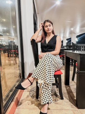 Siyah yelek ve puantiyeli şık bir kadın, geniş pencereli ve zarif dekorlu modern bir restoranda güven içinde poz veriyor. Moda, yaşam tarzı ve yemek temaları için mükemmel. Yüksek kalite, şık.