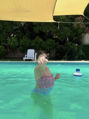 Renkli bir mayo giymiş genç bir kadın, parlak bir havuzda, sarı bir gölgelik ve yemyeşil bir arka planda, şakacı bir şekilde işaret ediyor. Yaz, yaşam tarzı ve tatil temalı projeler için mükemmel..