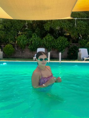 Renkli bir mayo giymiş genç bir kadın, parlak bir havuzda, sarı bir gölgelik ve yemyeşil bir arka planda, şakacı bir şekilde işaret ediyor. Yaz, yaşam tarzı ve tatil temalı projeler için mükemmel..